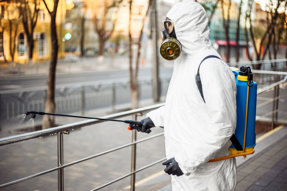 Man sprays disinfector onto the railing wearing coronavirus protective suit and equipment. Cleaning and sterilizing the not crowded city streets. Covid 19 nCov2019 spread prevention.